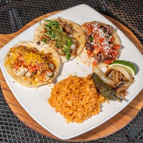 Three tacos with various toppings, rice, grilled onions, and a jalapeño on a white plate.