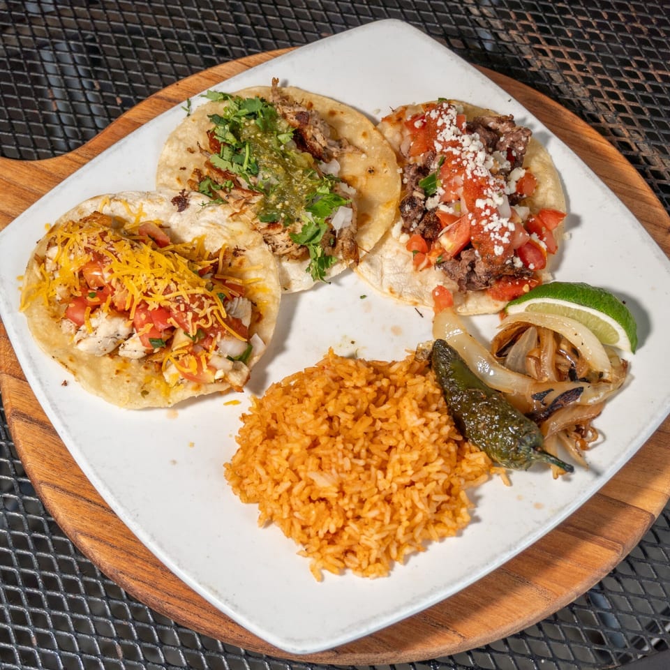 Three tacos with various toppings, rice, grilled onions, and a jalapeño on a white plate.