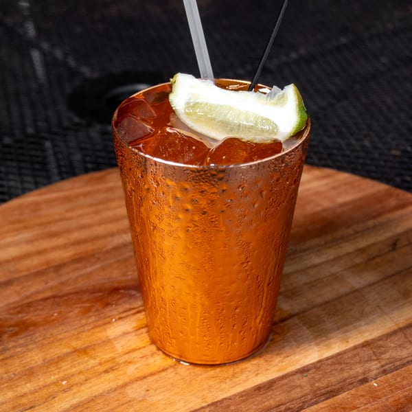 A cold Moscow Mule in a copper mug with a lime wedge, straws, and ice on a wooden table.