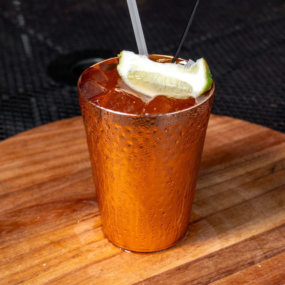 A cold Moscow Mule in a copper mug with a lime wedge, straws, and ice on a wooden table.