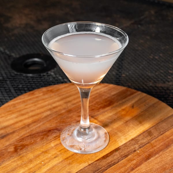 A light pink cocktail in a martini glass sits on a wooden board.