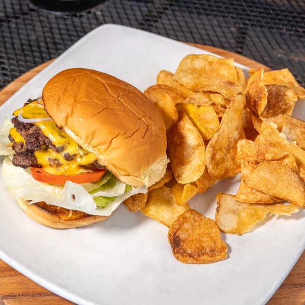 Double cheeseburger with lettuce, tomato, onion, and a side of crispy potato chips on a white plate.