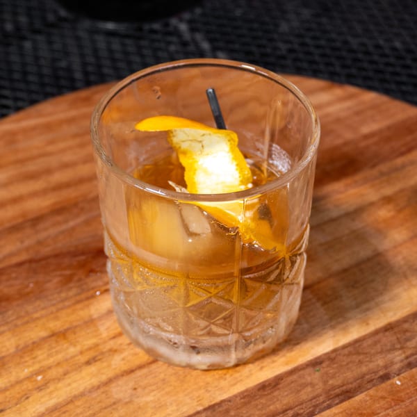 An Old Fashioned cocktail with a large ice cube and orange peel garnish on a wooden surface.