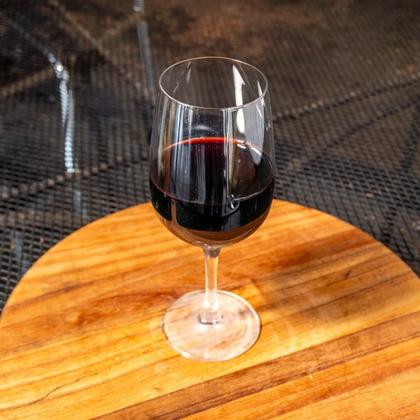 A glass of red wine sits on a round wooden board with a dark, textured background.