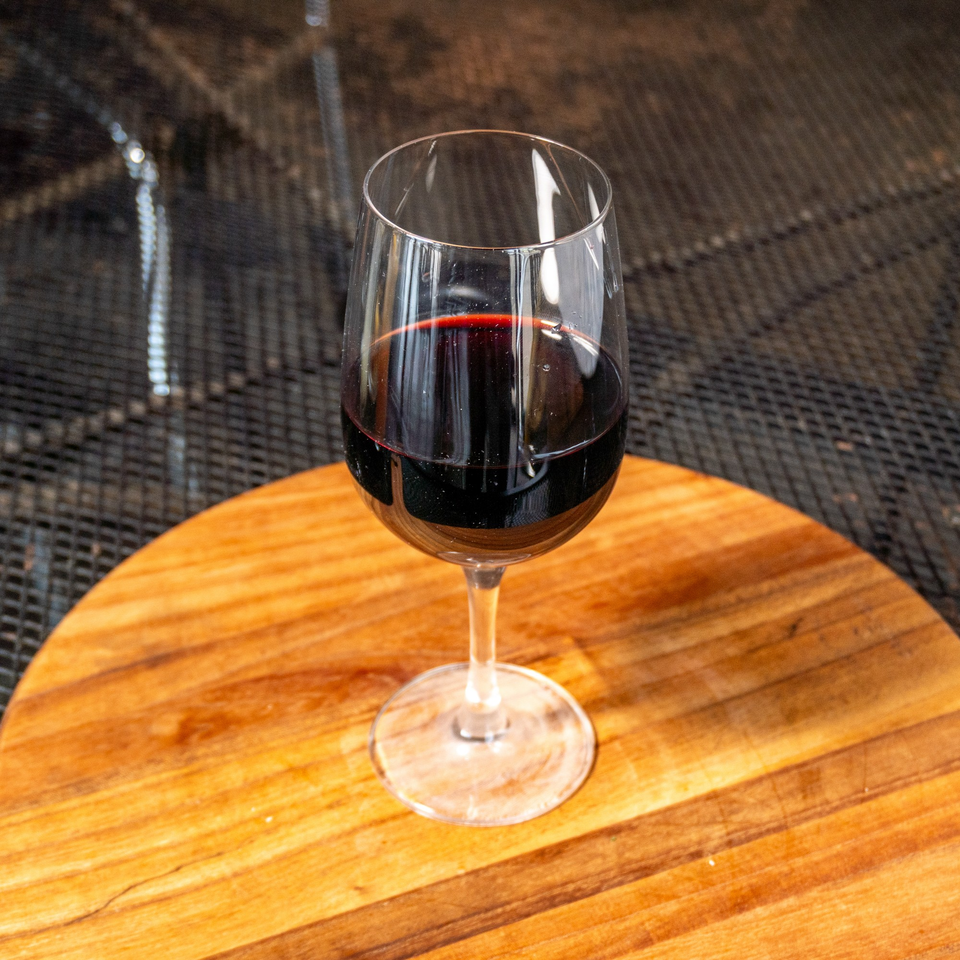 A glass of red wine sits on a round wooden board with a dark, textured background.