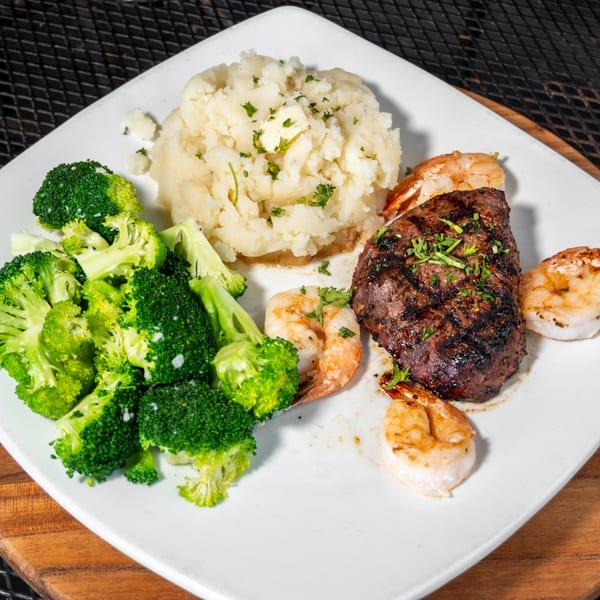 Grilled steak and shrimp with mashed potatoes and broccoli on a white plate.