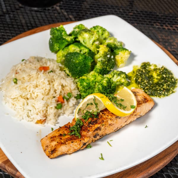 Grilled salmon with lemon slices, rice pilaf, steamed broccoli, and green pesto on a white plate.